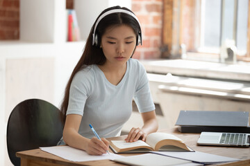 Concentrated Asian female student wearing earphones studying with textbooks making notes in...