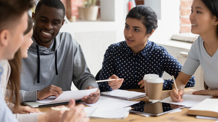 Multiracial diverse students sit at shared desk talk work with paperwork discuss work on group...
