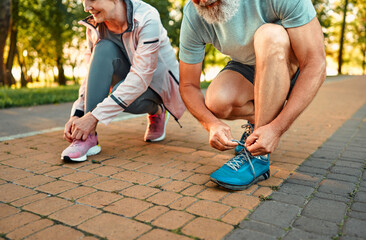 Active retirement concept. Close up of caucasian senior man and woman in active clothes tying laces on sport sneakers at city park. Healthy married couple preparing for morning run during summer day. - obrazy, fototapety, plakaty