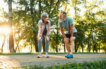 Recreation after workout. Happy old couple leaning with hands on knees and looking at each other while resting after outdoors jogging. Married man and woman taking care of health by common training. - obrazy, fototapety, plakaty