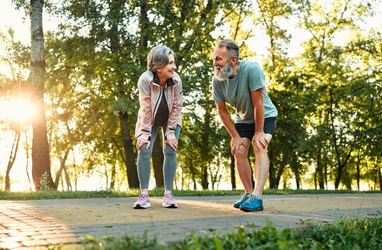 Recreation after workout. Happy old couple leaning with hands on knees and looking at each other while resting after outdoors jogging. Married man and woman taking care of health by common training.