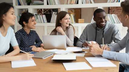 Overjoyed multiracial young people sit at table with laptop and paperwork have fun studying together, smiling happy multiethnic diverse millennial students laugh preparing for test brainstorming