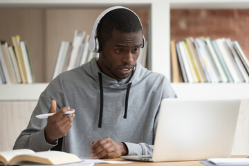 Frustrated african American millennial guy in headphones make notes studying using laptop, confused...