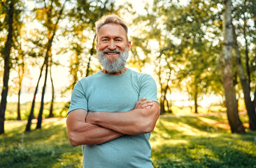 Healthy retirement lifestyle. Muscular grey bearded man keeping arms crossed and smiling while posing at green city park. Caucasian pensioner in sport clothes feeling happiness from active hobby.