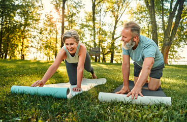 Yoga practice on fresh air. Sporty aged man and woman in active attire chatting and smiling while putting two mats on grass during sunny morning weather. Concept of active and healthy retirement.