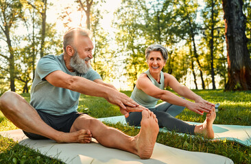 Flexible exercises for body. Sporty man and woman with grey hair stretching on yoga mats with hands to one leg during outdoors workout. Happy married couple with bare feet warming up together at park. - Powered by Adobe