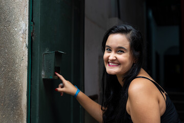 Portrait of beautiful young woman with black hair ringing the doorbell of a house and smiling.