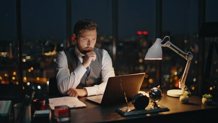 Late businessman searching data typing laptop at night cityscape in office.