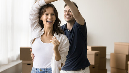 Cheerful young couple enjoying moving into new house, dancing in empty room with cardboard...