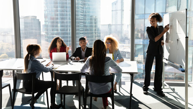 Business education. Diverse group of young mixed race employees gathered in boardroom on training workshop. Corporate team sit at conference table listen to black woman leader presenter speak by board