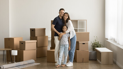 Happy parents and pretty kid girl moving into new house full length wide portrait. Young mom and...