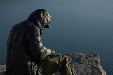 Man sitting on rocks overlooking Lake Titicaca with book in hand and cell phone on his right leg