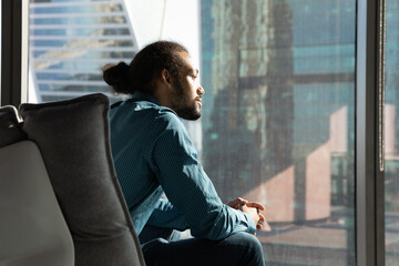 Creating idea. Pensive biracial businessman sit in cozy ergonomic armchair by window of corporate office look at distance. Young black guy team leader think on business vision strategy plan. Copyspace - Powered by Adobe