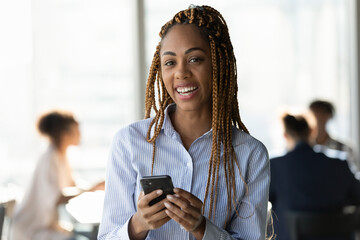 Modern day office worker. Headshot portrait of casual millennial biracial woman with stylish cornrows look at camera hold smartphone in hands. Young lady student employee networking online using phone