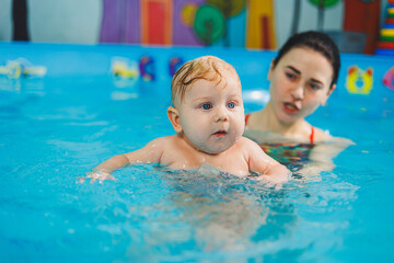 Fototapeta na wymiar A female trainer teaches a baby to swim in the pool and supports his hands. Baby swimming in the pool. Teaching a newborn boy to swim in the pool