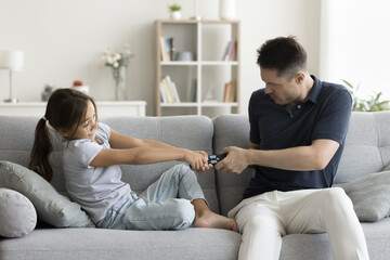 Serious stubborn father and naughty little kid fighting for remote control on couch, arguing about...