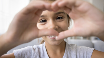 Happy cute little school kid girl looking at camera through hand heart frame. Positive pretty child joining fingers in gesture of friendship, love, happiness, enjoying childhood. Banner shot