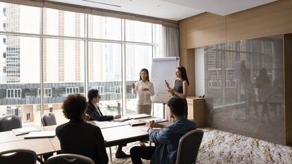 Two diverse speaker women presenting marketing report on whiteboard to coworkers, managers,...
