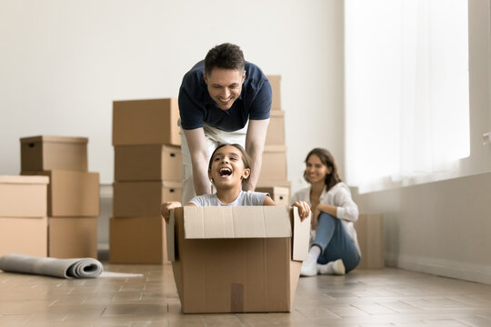 Happy young family having fun, moving into new flat. Cheerful dad walking and pushing forward paper box with excited screaming kid girl inside, laughing, celebrating successful mortgage
