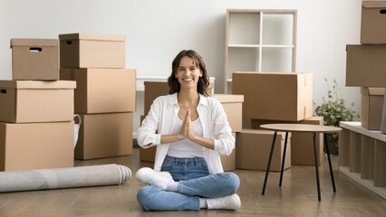 Happy young yogi girl meditating among moving boxes, relaxing after relocation, transportation...