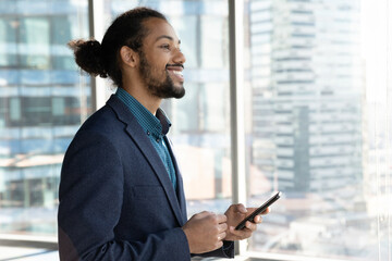 Smiling biracial employee admire cityscape from panoramic office window hold phone think on business achievement. Dreamy young black man corporate worker enjoy good news of project success. Copy space