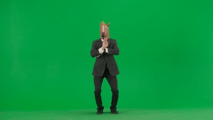 A man in a business suit with a horse head mask on a green studio background. Businessman dancing merrily. Hard office work concept.