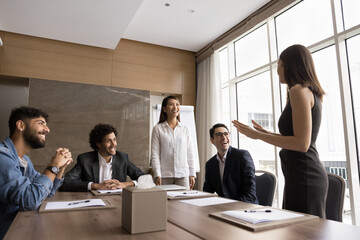 Cheerful multiethnic business professional colleagues working on project together, discussing creative ideas, brainstorming, standing and sitting at meeting table. Leader appraising Asian employee