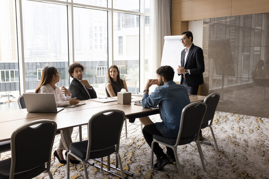 Multiethnic business team and project manager meeting in modern city office interior with large window, talking at conference table, discussing work strategy, brainstorming, listening to leader