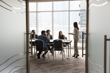 Candid shot through doorway of diverse business team and female leader discussing teamwork at large table. Group of young entrepreneurs negotiating, networking in modern office meeting room - obrazy, fototapety, plakaty