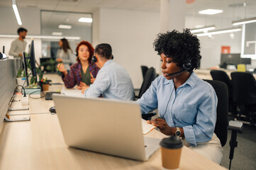 African american woman with headset working on a laptop in an open space office - obrazy, fototapety, plakaty