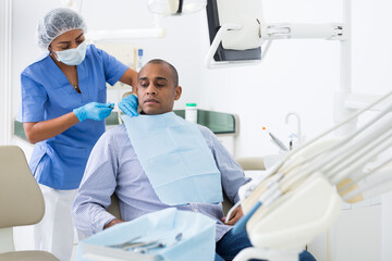 Obraz premium Portrait of adult hispanic man sitting on dental chair while woman dentist preparing him for medical examination in clinic