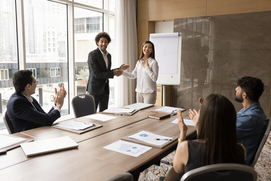 Cheerful young Arab team leader giving handshake to successful Asian professional girl, congratulating on job success, achievement. Multiethnic team applauding happy coworker on meeting