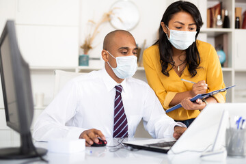 Portrait of two office employees in medical masks concentrating on work with papers and laptop....