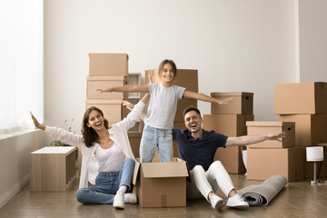 Happy little kid girl dancing in cardboard box, standing in paper container with open hands near cheerful mom and dad sitting on floor. Young couple and child enjoying moving activity