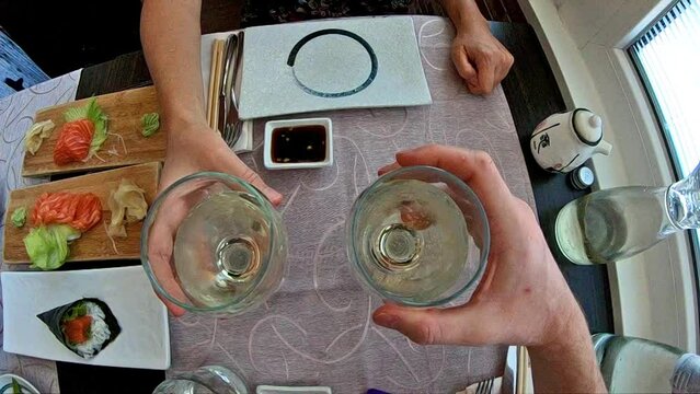 SLOW MOTION: Aerial view of two white wine glasses while doing cheers to celebrate an event in a japanese sushi restaurant interior, set table background and slow-motion effect.