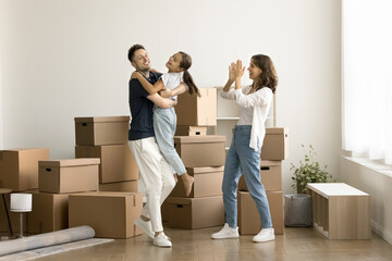 Fototapeta na wymiar Happy joyful parents and kid girl moving into new comfortable house, apartment, flat, celebrating property buying, successful mortgage for young family, dancing at stacked boxes