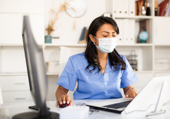 Hispanic woman general practitioner wearing medical mask working with case histories on laptop in...