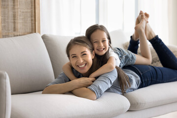 Little girl lying on moms back, smile look together at camera. Young woman enjoy carefree leisure lay on couch in cozy living room stare at cam feel happy. Custody, ties, motherhood, family relaxation