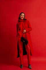 Fashionable happy smiling woman wearing trendy red midi coat, turtleneck sweater, leather pants, zebra print boots, holding baguette bag, posing on red background. Full-length studio fashion portrait