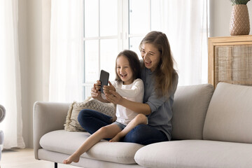 Overjoyed family relax sitting on sofa in living room, loving mother show funny mobile app to...