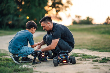 Father and son fixing a remote control toy car on a dirt road in nature.