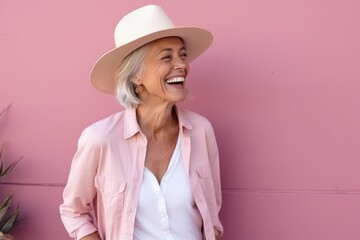 Portrait of happy senior woman in hat standing against pink wall.
