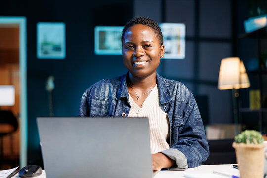 Black Woman Working At Her Laptop In A Modern Apartment, Studying Online And Researching Information For Her Project. Smiling African American Blogger Using Personal Computer While Looking At Camera.