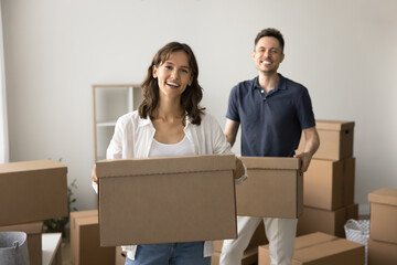 Happy young couple carrying moving boxes in new apartment, house, walking past heap of paper containers, looking at camera, smiling, enjoying relocation activities, beginning living together