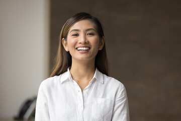 Happy beautiful young Asian manager woman in white formal shirt posing indoors, looking at camera, smiling, showing white perfect teeth. Successful female business leader head shot portrait