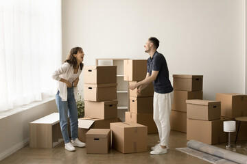 Cheerful young couple celebrating new flat buying, standing at heap of stacked cardboard boxes, talking, laughing, discussing successful mortgage, moving into new apartment, flat