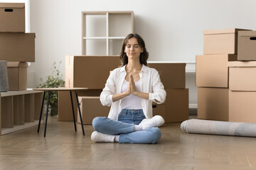 Peaceful young home owner woman meditating during stressful moving into new home, sitting on floor,...