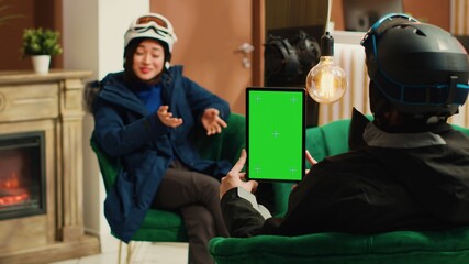 Traveller shows isolated greenscreen on tablet while she sits with skiing partner in lounge area,...