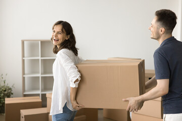 Joyful young married couple carrying large moving box, walking in row past stacked paper containers...