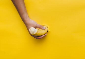 A man's hand holds a penis-shaped banana from a torn hole in yellow paper. Comic concept of masturbation, erection and arousal.